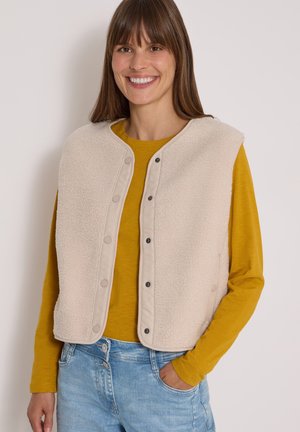 Kvinde smiler iført en beige vest med trykknapper over en sennepsfarvet langærmet skjorte og lyseblå jeans med den ene hånd i lommen.