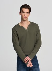 Długorękawowa koszulka Henley w oliwkowej zieleni, o prążkowanej fakturze, z trzema guzikami, dopasowany krój, zestawiona z ciemnymi dżinsami.