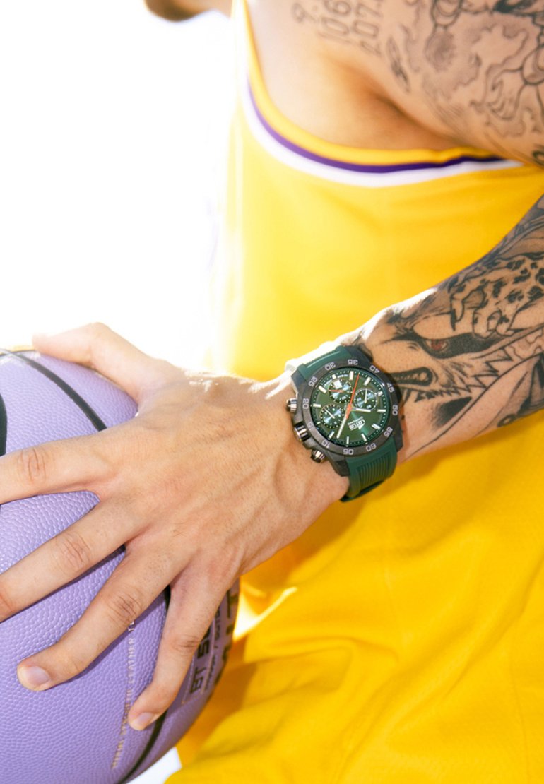 Reloj verde con pantalla digital y analógica, acentos negros y una correa texturizada. Una mano sostiene un balón de baloncesto morado, llevando una camiseta deportiva amarilla.