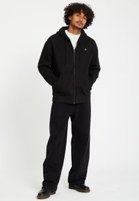 Black zip-up hoodie with a kangaroo pocket, drawstring hood, and logo accent. Paired with loose-fitting black pants and black shoes.