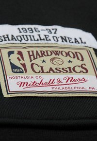 Schwarzer Stoff mit einem gewebten Etikett, das "1996-97 SHAQUILLE O'NEAL", "HARDWOOD CLASSICS" und "Mitchell & Ness" in Rot und Creme zeigt.
