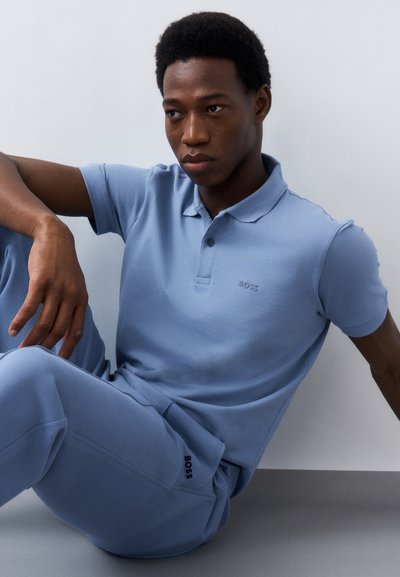 Homme portant un polo bleu clair et un pantalon de survêtement avec le logo BOSS, assis contre un mur blanc uni et un sol gris.