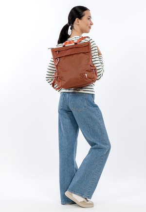 Brown textured backpack with adjustable straps and multiple zip pockets. Worn over a striped long-sleeve shirt and paired with wide-leg jeans.