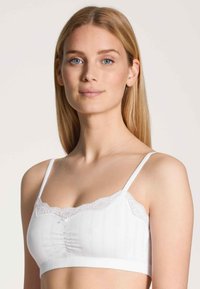 Witte bralette met kantafwerking bij de halslijn, geplooid middenstuk en verstelbare bandjes. Glad materiaal en gevoerd band voor comfort en ondersteuning.