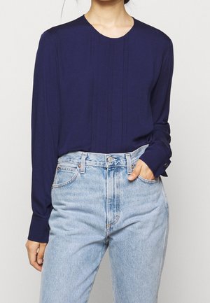 Blusa lunga blu navy con colletto rotondo e pieghe frontali. Abbinata a jeans in denim chiaro a vita alta. Tessuto morbido e liscio.