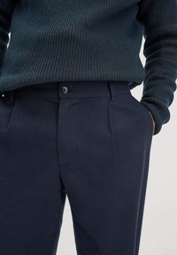 Donkerblauwe broek met een gladde textuur, voorzien van een enkele knoopsluiting en zijzakken, gedragen met een donker geribbelde trui.