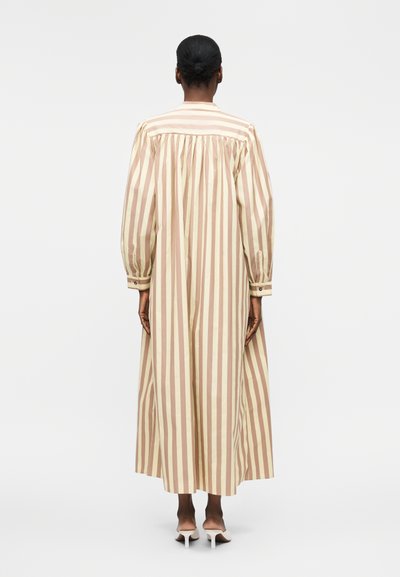 Γυναίκα με μαλλιά σε κότσο, φορώντας μια μακριά φόρεμα με κατακόρυφες ρίγες σε μπεζ και ανοιχτό καφέ και λευκά παπούτσια με τακούνια, στέκεται με την πλάτη γυρισμένη.