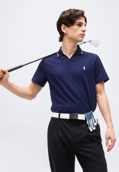 Νέος άνδρας με μπλε πόλο και μαύρο παντελόνι κρατάει ένα μπαστούνι του γκολφ στον ώμο του με ένα γάντι γκολφ κρυμμένο στη λευκή του ζώνη.