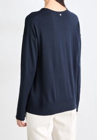 Osoba ubrana w granatowy sweter z długim rękawem i prążkowanym wykończeniem oraz białe spodnie, widoczna z tyłu na jednolitym tle.