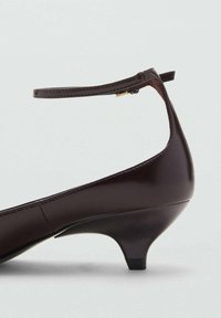 Zapato de tacón bajo de cuero marrón oscuro con una delgada correa en el tobillo y un pequeño hebilla dorada, exhibido sobre un fondo claro.