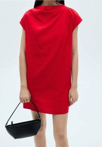 Rotes Kleid mit kurzen Ärmeln und lockerem Sitz, mit einem drapierten Ausschnitt. Modell hält eine kleine schwarze Tasche mit geschwungener Form und Tragegurt.