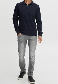 Polo à manches longues bleu marine avec un col zippé, poignets côtelés, porté avec un jean gris clair et des baskets noires à semelles blanches.