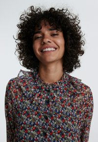 Blumenshirt mit Rüsche am Kragen, transparentem Stoff und einer Mischung aus roten, blauen und gelben Blumen; mit Knopfleiste vorne.