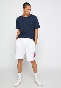 Camiseta de algodón azul marino de mangas cortas, combinada con pantalones cortos de algodón blancos que presentan un estampado gráfico rojo. Completa con zapatillas blancas y calcetines.