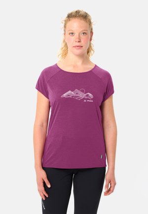 Femme aux cheveux blonds bouclés portant un t-shirt violet à manches courtes avec un motif de lignes de montagnes et un pantalon noir, debout devant un fond uni.