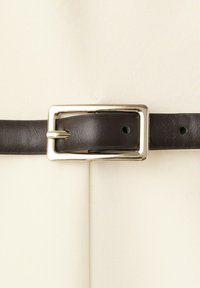 Tmavě hnědý kožený pásek provlečený obdélníkovou stříbrnou kovovou přezkou, zapnutý přes hladkou krémovou látku.