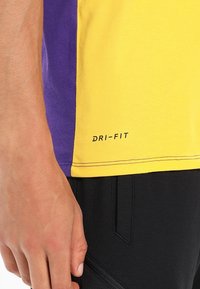 T-shirt à manches courtes Dri-FIT avec sections jaunes et violettes, présentant une texture lisse et un logo noir imprimé près de l'ourlet.