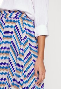 Pleated hame, jossa on geometrinen sahalaitakuosi sinisellä, violetilla, ruskealla ja valkoisella. Valmistettu kevyestä kankaasta, joka tuntuu sileältä.