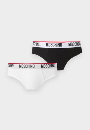 Uomo muscoloso in piedi a piedi nudi che indossa slip neri con fascia bianca con la scritta "Moschino" su sfondo bianco uniforme.