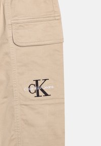 Béžové cargo kalhoty s bočními kapsami, obsahující vyšívané logo "Ck" v černé barvě a "Calvin Klein Jeans" v bílém stehu.