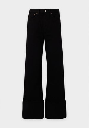 Femme debout portant un haut rose ajusté à manches courtes, un pantalon noir large taille haute avec revers roulés, et des chaussures blanches pointues.