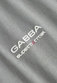 Šedá tkanina s hladkým povrchom, na ktorej sa nachádza biele vyšívané logo "GABBA supercortical." Logo je umiestnené na prominentnom mieste v strede.