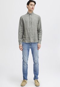 Camicia a maniche lunghe in lino grigio con colletto button-down, chiusura a bottoni e logo discreto. Abbinata a jeans azzurri e scarpe beige.