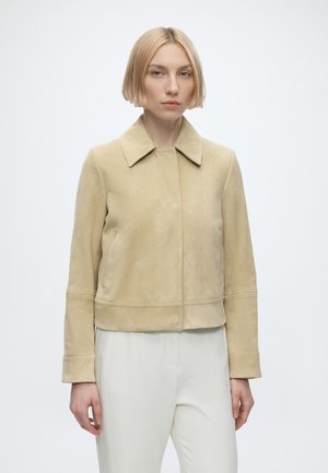 Kvinde med kort blondt hår iført en beige ruskindsjakke og hvide bukser, stående mod en ensfarvet lys baggrund.