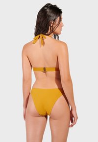 Žuti bikini s halter vratom i detaljem za vezivanje, sadrži pravokutnu kopču i visoko rezane strane. Glatka tkanina i minimalno šivanje.