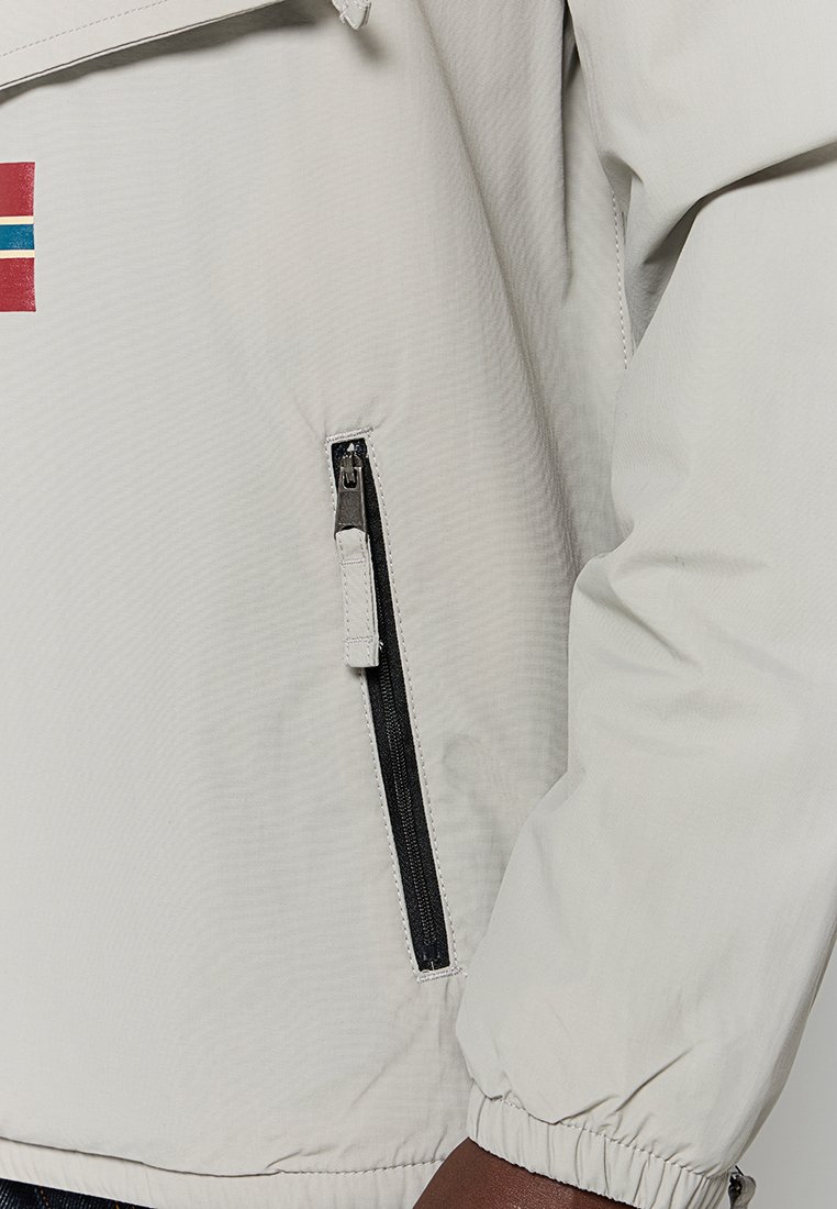 Veste gris clair avec une poche zippée, des poignets élastiques et un détail de bande horizontale colorée sur la poitrine. Tissu texturé, résistant au vent.