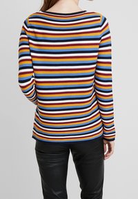 Haut à manches longues à rayures multicolores horizontales en rouge, jaune, bleu et blanc sur un fond sombre. Tissu doux et texturé.