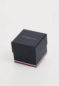 Dunkelblauem quadratischer Karton mit einem Deckel, der weißen Text "TOMMY HILFIGER UHREN" und einem rot-weiß-blauen gestreiften Akzent an der Basis aufweist.