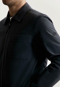 Giacca nera realizzata in un materiale leggero e liscio. Presenta un colletto a punta, una zip frontale e una tasca sul petto. Design minimalista con dettagli del logo.
