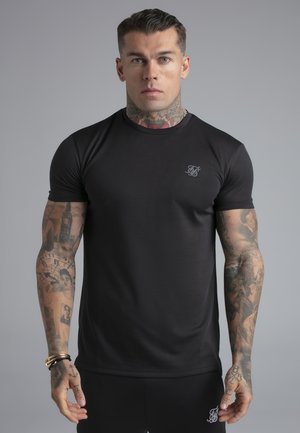 Mężczyzna z tatuażami, ubrany w czarną koszulkę z krótkim rękawem i szorty, stojący na jednolitym szarym tle o neutralnym wyrazie twarzy.