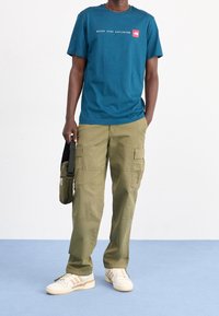 Camiseta de algodón color azul verdoso con el texto "NUNCA DEJES DE EXPLORAR", pantalones cargo oliva con bolsillos laterales y zapatillas color crema con detalles en marrón.