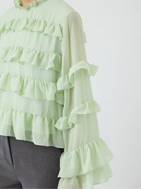 Světle zelená blůza s volánovými vrstvami, dlouhými rukávy a vysokým výstřihem. Průsvitný materiál s jemnou texturou. V kombinaci se šedými kalhotami.