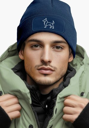 Navy knit beanie with a textured pattern and a logo patch featuring a dog silhouette. Worn with a green jacket and black inner layer.