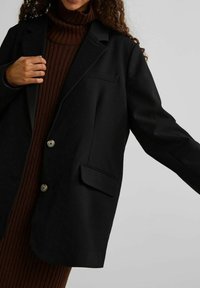 Schwarzer übergroßer Blazer aus strukturiertem Stoff, mit zwei vorderen Knöpfen, einer Brusttasche und einem Reverskragen, kombiniert mit einem braunen gerippten Rollkragenpullover.