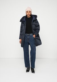 Navy puffer coat with a hood, black sweater, and baggy jeans. Model wears a light pink beanie and black heeled boots. Smooth texture throughout.