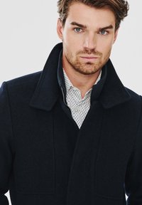 Ciemnoniebieski wełniany płaszcz z dużym kołnierzem i detalami kieszeni, nałożony na wzorzystą białą koszulę. Gładka faktura i strukturalny krój.