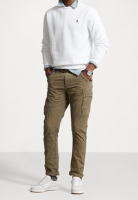 Felpa bianca in cotone, camicia in denim chiaro sotto, e pantaloni cargo olive con tasche multiple. Sneakers bianche completano l'outfit.