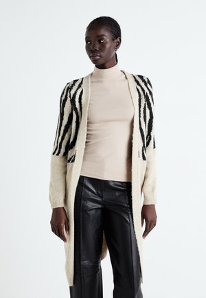 Zebra-Muster Strickjacke in Creme und Schwarz, gestrickte Textur, offene Vorderseite, lange Ärmel, kombiniert mit einem beigen Top und schwarzen Lederhosen.