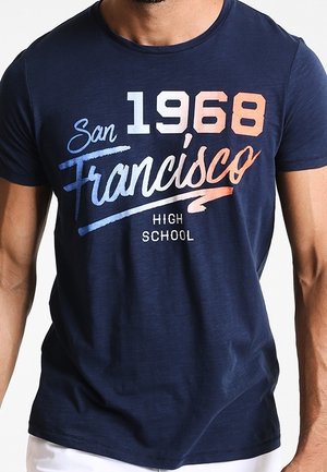 Mężczyzna noszący granatową koszulkę z napisem „San Francisco 1968 High School” w niebieskich, białych i pomarańczowych literach.