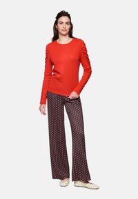 Red long-sleeve top with gathered detail on sleeves, paired with dark patterned wide-leg pants featuring a geometric design. Light-colored footwear.