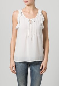Blusa bianca senza maniche con dettagli arricciati sulle spalle, fronte plissettato e chiusura con fiocco. Tessuto leggero e semi-trasparente.
