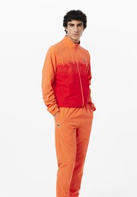 Completo sportivo in sfumature di arancione, composto da una giacca con zip e pantaloni coordinati. Entrambi i pezzi hanno una finitura testurizzata e dettagli con logo verde.