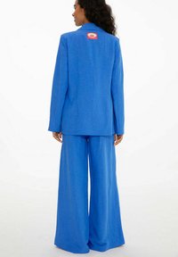 Modrá sako a kalhoty s širokými nohavicemi, vyrobené z měkkého materiálu. Sako má na zádech červenou nášivku s logem. Jednoduchý design, přiléhavý střih.