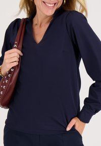 Glunderende vrouw in een marineblauwe blouse met V-hals, houdt met één hand een bruine schoudertas met studs vast, andere hand in haar broekzak.