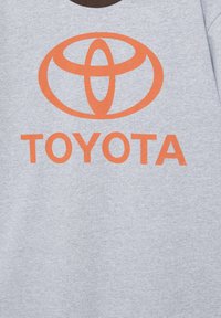 Sivé bavlnené tričko s oranžovým logom Toyota a nápisom. Golier je hnedý, s okrúhlym strihom. Materiál má jemnú textúru.