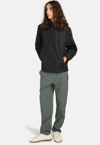 Giovane con lunghi capelli ricci, indossa una giacca nera con zip, pantaloni verdi e sneakers bianche, in piedi con le mani nelle tasche.
